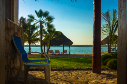 Tropical waterfront patio with palm trees, thatched roof shade, lounge chairs, and water view